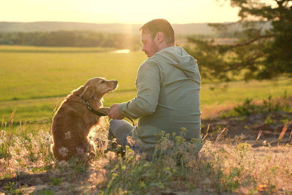 A man in a light jacket sits on the grass with a golden retriever at sunset. They are looking at each other, and the dog is holding a leash in its mouth. The background features a scenic view with trees and open fields.