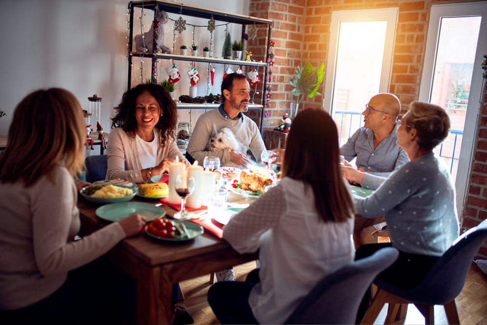 A family gathers around a festive dining table, enjoying a meal together. The room is decorated with holiday ornaments, and a small dog sits on one person's lap. Warm light illuminates the scene, creating a cozy atmosphere.